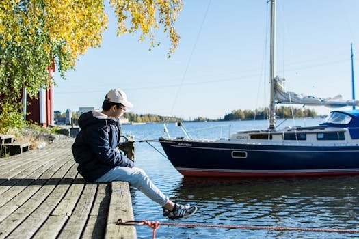 A young person sitting by a Finnish lake with a sailboat in the background during a sunny day.