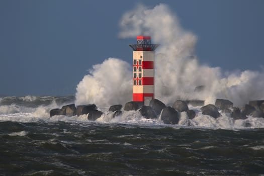 Red and white lighthouse with waves crashing against rocks under a stormy sky.