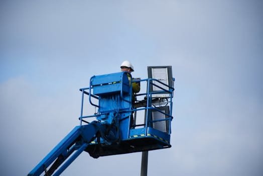 An industrial worker wearing a hard hat operating a lift platform for maintenance tasks in an outdoor setting.