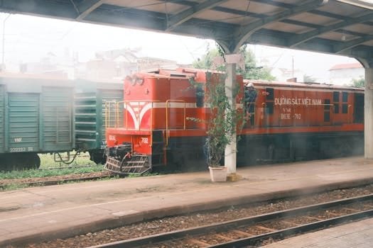 A red train parked at a Vietnamese urban station under a covered platform. Industrial and urban feel.