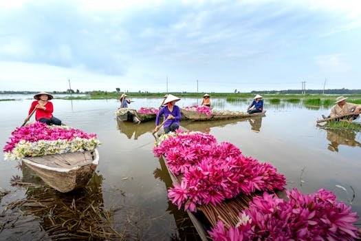 Women in conical hats paddling boats filled with flowers on a serene river during the day.