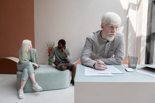 Albino worker standing at table and writing on paper in office with African American man sitting with woman on background