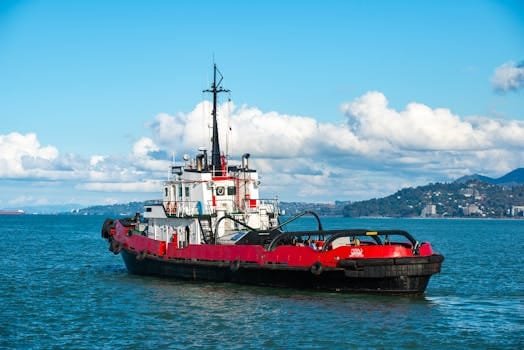 A vibrant red tugboat navigates the calm blue sea under a clear sky with clouds.