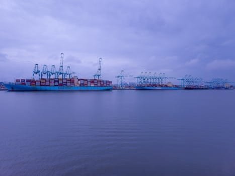Container ships docked with cranes at an industrial port under a cloudy sky.