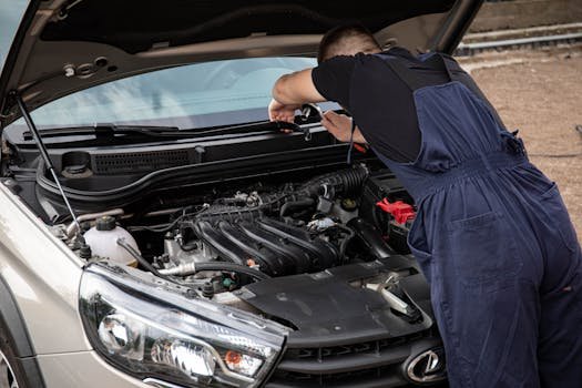 A mechanic in blue overalls works on a car engine in an outdoor setting.