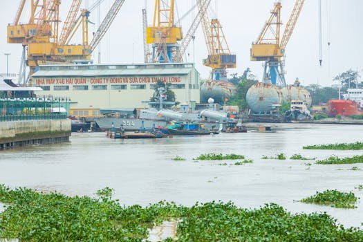 A bustling Vietnamese shipyard with cranes, military ships, and vibrant greenery.