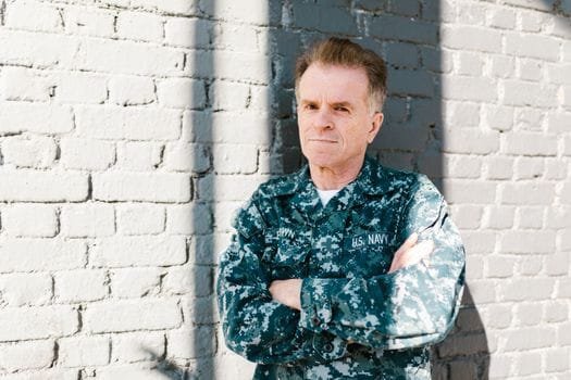 Cum să Alegi Cursul Ofițer Punte Diploma Acreditată: Compararea Ofertei Educaționale 4 Senior Navy veteran standing proudly in naval uniform against a brick wall, outdoors in daylight.