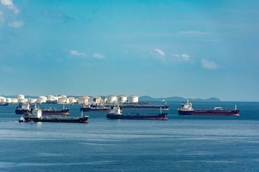 A fleet of cargo ships docked near oil storage tanks along a serene coastline with a clear blue sky above.