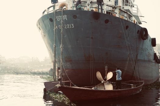 A worker on a small boat beside a large docked ship in Dhaka, Bangladesh.