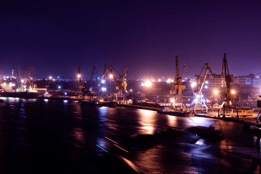 Curs MARPOL vs. Curs GMDSS: Ce Alegi pentru Cariera ca Ofițer Maritim în România? 4 A serene night view of illuminated shipping cranes at a bustling harbor, reflecting lights over calm water.