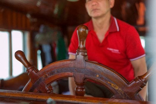 Top 5 Cursuri Certificare GMDSS Maritim pentru Frecvențe Radio: Alegerea Ideală 3 Close-up of a man in red shirt at a ship's wooden helm, steering indoors.
