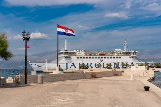 Jadrolinija ferry at Supetar harbor with Croatian flag under a clear sky.
