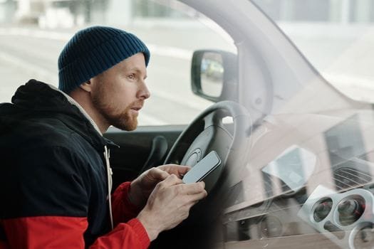 Caucasian man in a knitted cap using a smartphone while sitting in a parked car.