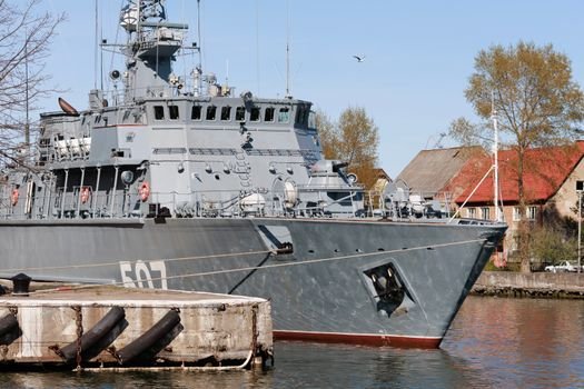 A military ship docked at a harbor with structures and trees in the background under clear skies.
