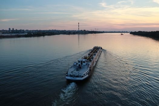 A cargo ship travels on a calm river at sunset with a city skyline in the background.