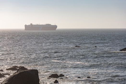 A cargo ship sails on a calm ocean under a clear sky, viewed from a rocky shore.
