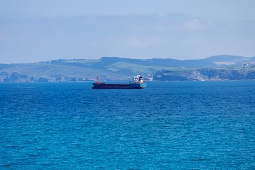 A lone cargo ship navigating serene blue waters with a distant coastline in view under a clear sky.