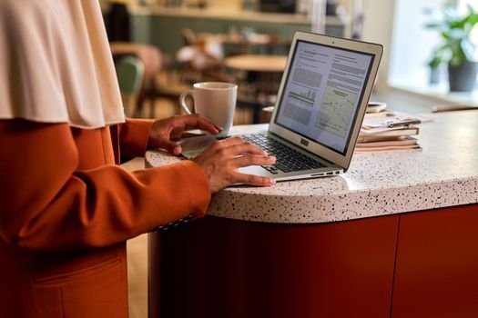 Adult woman typing on a laptop at a modern office counter, coffee beside her in a casual indoor setting.
