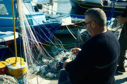 Elderly man repairing fishing net at a harbor with boats in the background.