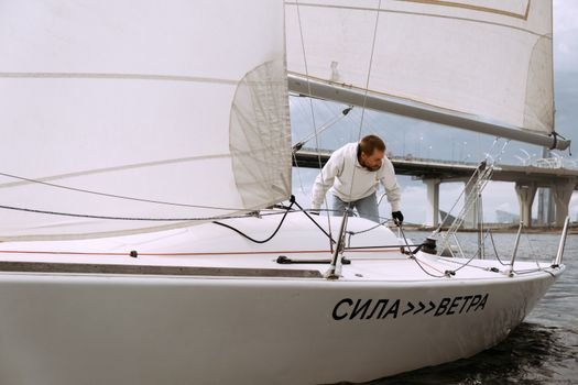 Joburi Maritime Vara 2026 vs Pe Tot Parcursul Anului: Ce Aleg Pentru Marinarii Români? 2 Man navigating yacht on the water near a city bridge, showcasing nautical adventure and sailing skills.