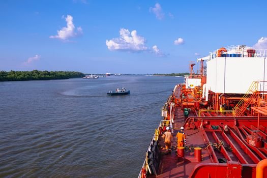 A cargo ship sails along a wide river with crew members visible on deck under a clear blue sky.