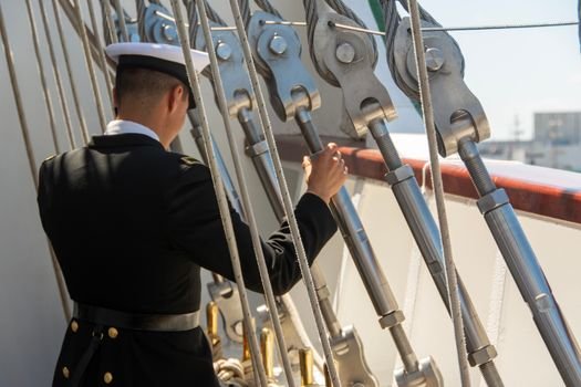 A sailor in uniform adjusts ropes on a ship deck during the day.
